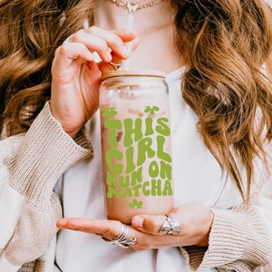 May include: A glass tumbler with a bamboo lid and a green graphic that says "This Girl Runs on Matcha". The tumbler is filled with a brown liquid and a straw is sticking out of the top.