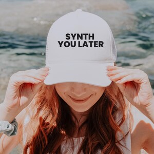 May include: A white mesh baseball cap with a black embroidered text that reads "SYNTH YOU LATER". The cap is worn by a person with long, wavy, red hair.