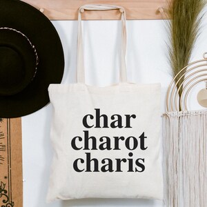 May include: Cream-colored tote bag with the words "char charot charis" printed in black. A black hat hangs on the left, and a decorative wall hanging is on the right. A shelf above holds a small potted plant and other decorative items.