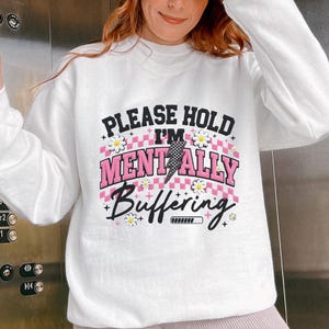 Puede incluir: Sudadera blanca con el texto "PLEASE HOLD I'M MENTALLY BUFFERING" en negro y rosa. El diseño incluye detalles de margaritas y una barra de carga. La sudadera se combina con unos pantalones cortos de ciclista rosa claro.