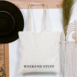 Può includere: Una borsa tote color crema con la scritta "WEEKEND STUFF" in nero. Sono visibili anche un cappello nero e un arazzo decorativo. Una mensola di legno sopra contiene una piccola pianta in vaso e altri oggetti decorativi.