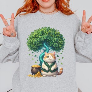 Puede incluir: Sudadera gris claro con un diseño del Día de San Patricio. El gráfico incluye un gato guiñando un ojo con un chaleco verde, un árbol hecho de tréboles, una olla de oro y tréboles dispersos. El gato es naranja y blanco.