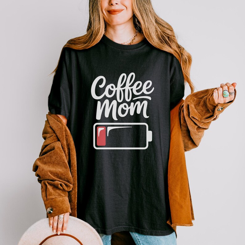 Flatlay photograph of a folded dark navy blue vintage-wash t-shirt surrounded by black and white sneakers, blue jeans, and a woven net bag. The shirt features the text "Coffee Mom" in white cursive and print, stacked above a graphic of a low battery symbol with a single red bar.