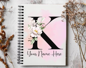 Könnte beinhalten: Ein weißes Spiralnotizbuch mit einem rosafarbenen Aquarellhintergrund und einem schwarzen Buchstaben K, der mit weißen Blumen verziert ist. Der Text "Your Name Here" ist unter dem Buchstaben gedruckt.