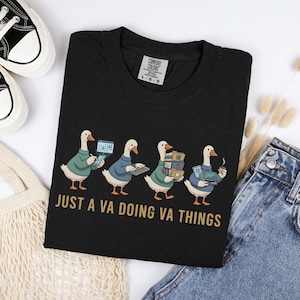May include: Black t-shirt featuring a graphic of four geese, each holding items like a laptop, keyboard, and files. The geese are wearing sweaters. The text "JUST A VA DOING VA THINGS" is printed below the geese in gold letters.