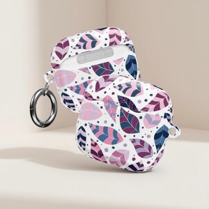 May include: White AirPod case with a colourful leaf pattern in shades of purple, pink, and blue. The case is open, revealing the inside. A silver metal ring is attached to the case.
