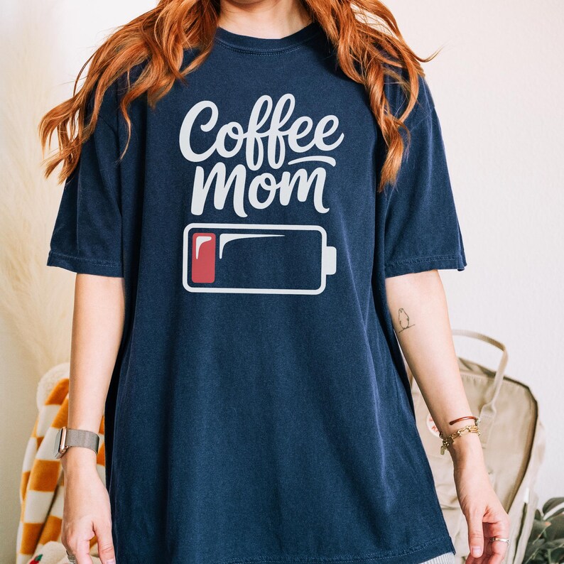Flatlay photograph of a folded dark navy blue vintage-wash t-shirt surrounded by black and white sneakers, blue jeans, and a woven net bag. The shirt features the text "Coffee Mom" in white cursive and print, stacked above a graphic of a low battery symbol with a single red bar.