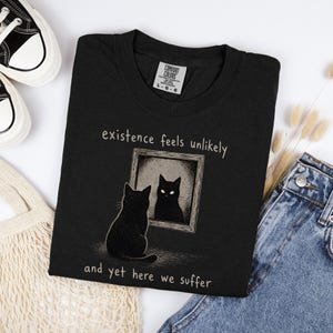 May include: A black t-shirt featuring a graphic of a black cat sitting in front of a framed mirror. The text above the mirror reads existence feels unlikely and the text below reads and yet here we suffer. The shirt has a Comfort Colors brand label.