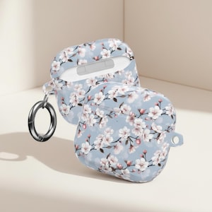 Könnte beinhalten: Zwei hellblaue AirPod-Hüllen mit einem Blumenmuster aus weißen und rosa Blüten. Eine Hülle ist geöffnet und gibt das Innere frei. Ein silberfarbener Metallring ist an der offenen Hülle befestigt.