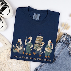 Könnte beinhalten: Ein marineblaues T-Shirt mit einer Grafik von vier weißen Enten, die als Krankenschwestern in Arbeitskleidung gekleidet sind. Die Enten führen medizinische Aufgaben aus, halten eine Spritze und tragen einen Stapel Akten. Der Text lautet "JUST A NARS DOING NARS THINGS."
