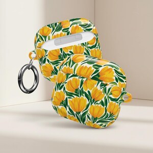 May include: Two AirPod cases with a white and yellow floral design. The cases feature a repeating pattern of yellow flowers and green leaves on a white background. One case is open, showing the white interior. A silver carabiner clip is attached.