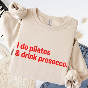 May include: A beige crewneck sweatshirt featuring the red text I do pilates & drink prosecco. printed on the front. The garment has long sleeves with ribbed cuffs and a ribbed neckline, showcasing a standard fit and a soft, solid-colored fabric.