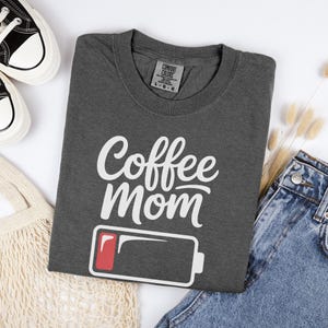 Flatlay photograph of a folded dark navy blue vintage-wash t-shirt surrounded by black and white sneakers, blue jeans, and a woven net bag. The shirt features the text "Coffee Mom" in white cursive and print, stacked above a graphic of a low battery symbol with a single red bar.