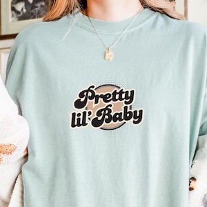 Könnte beinhalten: Hellblaues T-Shirt mit dem Text "Pretty lil' Baby" in einer Retro-Schriftart. Die Grafik ist in Schwarz, Braun und Weiß gehalten und vor einem kreisförmigen Hintergrund platziert. Das Shirt hat einen Rundhalsausschnitt und kurze Ärmel.