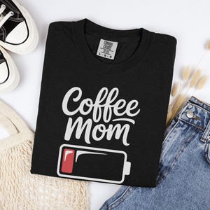 Flatlay photograph of a folded dark navy blue vintage-wash t-shirt surrounded by black and white sneakers, blue jeans, and a woven net bag. The shirt features the text "Coffee Mom" in white cursive and print, stacked above a graphic of a low battery symbol with a single red bar.