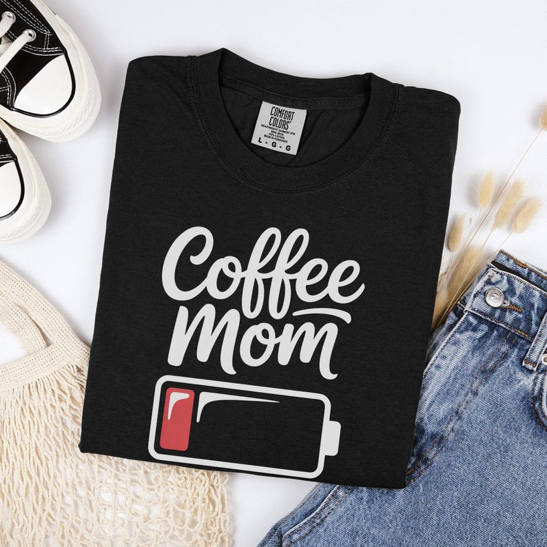 Flatlay photograph of a folded dark navy blue vintage-wash t-shirt surrounded by black and white sneakers, blue jeans, and a woven net bag. The shirt features the text "Coffee Mom" in white cursive and print, stacked above a graphic of a low battery symbol with a single red bar.