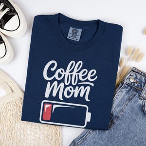 Flatlay photograph of a folded dark navy blue vintage-wash t-shirt surrounded by black and white sneakers, blue jeans, and a woven net bag. The shirt features the text "Coffee Mom" in white cursive and print, stacked above a graphic of a low battery symbol with a single red bar.