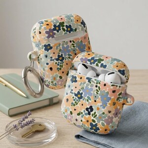May include: Two AirPod cases with a floral pattern, one open with white earbuds. The cases have a cream base with blue, orange, and purple flowers. One case has a silver carabiner clip attached for easy carrying.