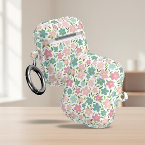 May include: Two white AirPod cases with a floral pattern in pink, blue, and green. The cases have a silver carabiner clip attached. The background is a light brown surface.
