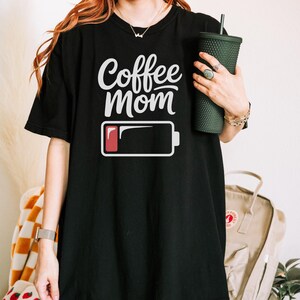 Flatlay photograph of a folded dark navy blue vintage-wash t-shirt surrounded by black and white sneakers, blue jeans, and a woven net bag. The shirt features the text "Coffee Mom" in white cursive and print, stacked above a graphic of a low battery symbol with a single red bar.