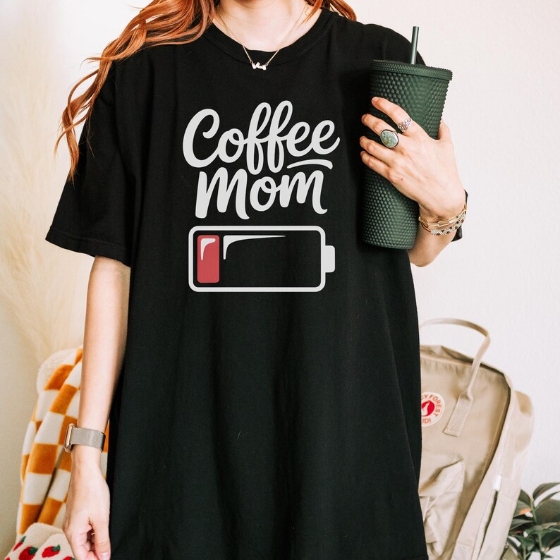Flatlay photograph of a folded dark navy blue vintage-wash t-shirt surrounded by black and white sneakers, blue jeans, and a woven net bag. The shirt features the text "Coffee Mom" in white cursive and print, stacked above a graphic of a low battery symbol with a single red bar.