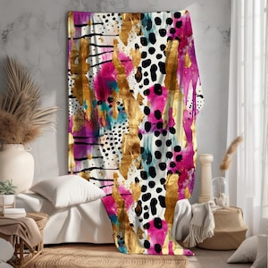 Könnte beinhalten: Eine dekorative Decke mit abstraktem Kunstmuster. Das Design zeigt goldene Pinselstriche, schwarze Punkte und Spritzer in Pink, Türkis und Weiß. Die Decke ist über ein Bett drapiert und zeigt ihr farbenfrohes, modernes Design.