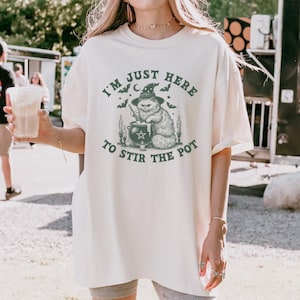 Puede incluir: Camiseta color crema con un gráfico de un gato con sombrero de bruja revolviendo un caldero. El texto "I'M JUST HERE TO STIR THE POT" está impreso encima y debajo de la ilustración del gato. La camiseta tiene un corte holgado.