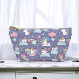May include: A lavender cosmetic bag with a repeating pattern of cartoon unicorns, clouds, and rainbows. The bag has a gold zipper and a rectangular shape, perfect for storing makeup or toiletries.