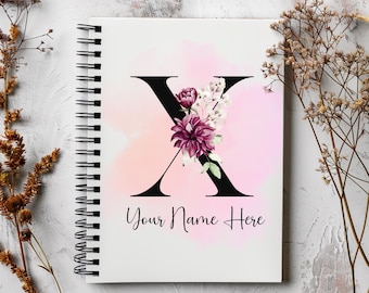 Könnte beinhalten: Ein weißes Notizbuch mit einer schwarzen Spiralbindung. Der Einband zeigt einen schwarzen Buchstaben X mit rosa und weißen Aquarellblumen. Der Text "Your Name Here" ist unter dem Buchstaben gedruckt.