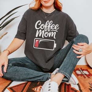 Flatlay photograph of a folded dark navy blue vintage-wash t-shirt surrounded by black and white sneakers, blue jeans, and a woven net bag. The shirt features the text "Coffee Mom" in white cursive and print, stacked above a graphic of a low battery symbol with a single red bar.