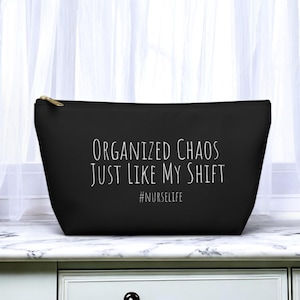 May include: Black zippered makeup bag with white text that reads "Organized Chaos Just Like My Shift #NURSELIFE"