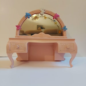 Könnte beinhalten: Ein rosa Spielzeug-Schminktisch mit Spiegel, verziert mit bunten Schmetterlingen und dekorativen Elementen. Der Schminktisch hat zwei Schubladen und geschwungene Beine, was eine verspielte Ästhetik erzeugt. Der Spiegel ist bogenförmig und hat einen dekorativen Rand.