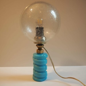Op de afbeelding: Een vintage tafellamp met een heldere, gespikkelde globe-kap en een turquoise, spiraalvormige voet. De lamp heeft messing accenten en een zichtbare gloeilamp. Het snoer is lichtbruin.