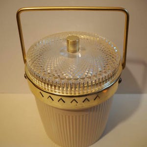 Op de afbeelding: Een vintage ijs emmer met een gouden handvat en een doorzichtig deksel met een diamantpatroon. De emmer is gemaakt van een crèmekleurig materiaal met verticale ribbels.