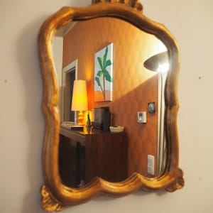 Op de afbeelding: Een wandspiegel met een sierlijk, geschulpt goudkleurig frame met een decoratieve kuif aan de boven- en onderkant. Het reflecterende glazen oppervlak toont een kamerinterieur met oranje muren, een lamp, een houten console en kunst aan de muur.