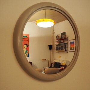Op de afbeelding: Een ronde spiegel met een grijs frame reflecteert een kamer met een gele en witte plafondlamp, een boekenkast met boeken en een stereo-installatie, en een schilderij van een vrouw in een kleurrijke jurk.
