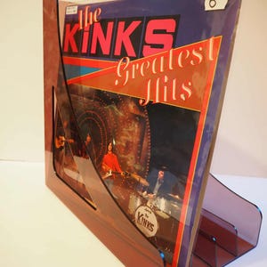 Op de afbeelding: Een transparante, hoekige vinyl recordhouder in een doorschijnende roodbruine kleur. De recordhoes toont de tekst "The Kinks Greatest Hits" in rood en wit, met een bandfoto. Een kleine witte sticker met het nummer 6 is zichtbaar.