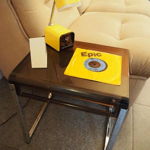 May include: A yellow desk lamp, a white rectangular object, and a vinyl record with the word "Epic" on the cover sit on a brown side table with chrome legs. The table is in front of a beige sofa.
