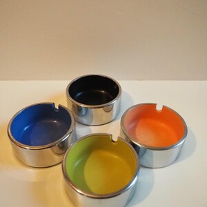Op de afbeelding: Een set van vier ronde asbakken met een gepolijste chromen buitenkant. Elke asbak heeft een gladde, effen gekleurde binnenkant in blauw, zwart, oranje of limoengroen en bevat een enkele kleine inkeping op de rand om voorwerpen op te leggen.