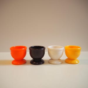 Op de afbeelding: Vier eierrekjes in verschillende kleuren: oranje, bruin, wit en geel. De eierrekjes zijn gemaakt van plastic en hebben een eenvoudig ontwerp.