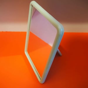 Può includere: Un piccolo specchio da toeletta rettangolare bianco sporco con angoli arrotondati. Lo specchio è appoggiato su un piccolo supporto, che riflette una tonalità rosata. La cornice ha una finitura morbida e opaca, su uno sfondo arancione.