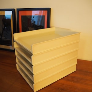 Op de afbeelding: Een stapel van vijf crèmekleurige, plastic brievenbakken. De bakken zijn rechthoekig met een lip aan de voor- en achterkant. De stapel staat op een houten oppervlak, met ingelijste kunstwerken op de achtergrond.
