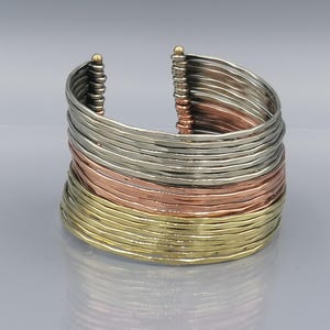 Könnte beinhalten: Ein mehrlagiges Armreif-Armband aus Silber, Kupfer und Gold. Das Armband besteht aus dünnen, gehämmerten Metallstreifen, die übereinander gestapelt sind.