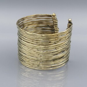 Könnte beinhalten: Ein goldfarbenes Metall-Armreif-Armband aus mehreren dünnen, wellenförmigen Bändern, die übereinander gestapelt sind. Das Armband hat ein schlichtes, minimalistisches Design.