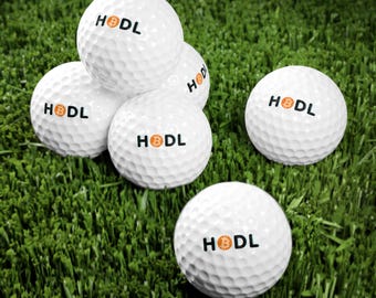 Pode incluir: Seis bolas de golfe brancas com a palavra "HODL" impressa em letras pretas. As bolas de golfe estão dispostas em forma de pirâmide sobre uma superfície verde coberta de grama.