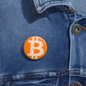 Könnte beinhalten: Nahaufnahme einer Jeansjacke mit einem runden, orangen Knopf, auf dem ein weißes Bitcoin-Symbol abgebildet ist.