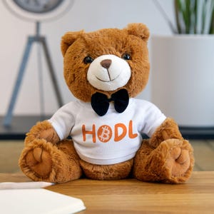 May include: A brown teddy bear wearing a white shirt with the word "HODL" and a Bitcoin symbol printed on it. The bear is also wearing a black bow tie.