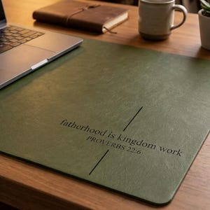 Könnte beinhalten: Eine grüne Schreibtischunterlage mit dem Text "fatherhood is kingdom work PROVERBS 22:6" in Schwarz. Ein Laptop, ein braunes Lederjournal, eine Tasse und eine kleine Topfpflanze stehen auf einem Holztisch.