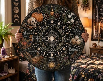 May include: A round, black decorative wall art piece featuring a seasonal wheel design. The wheel includes illustrations of a skull, pumpkin, eggs, and flowers, with text labels like "Mabon" and "Ostara". The design incorporates moon phases and botanical elements.