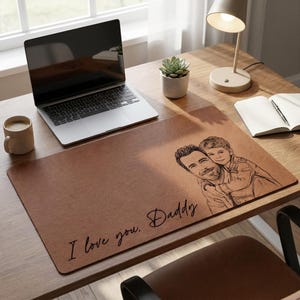 Op de afbeelding: Bruine leren bureauonderlegger met een schets van een vader en kind. De tekst "I love you, Daddy" staat op de mat. Een laptop, koffiemok en bureaulamp zijn ook zichtbaar.